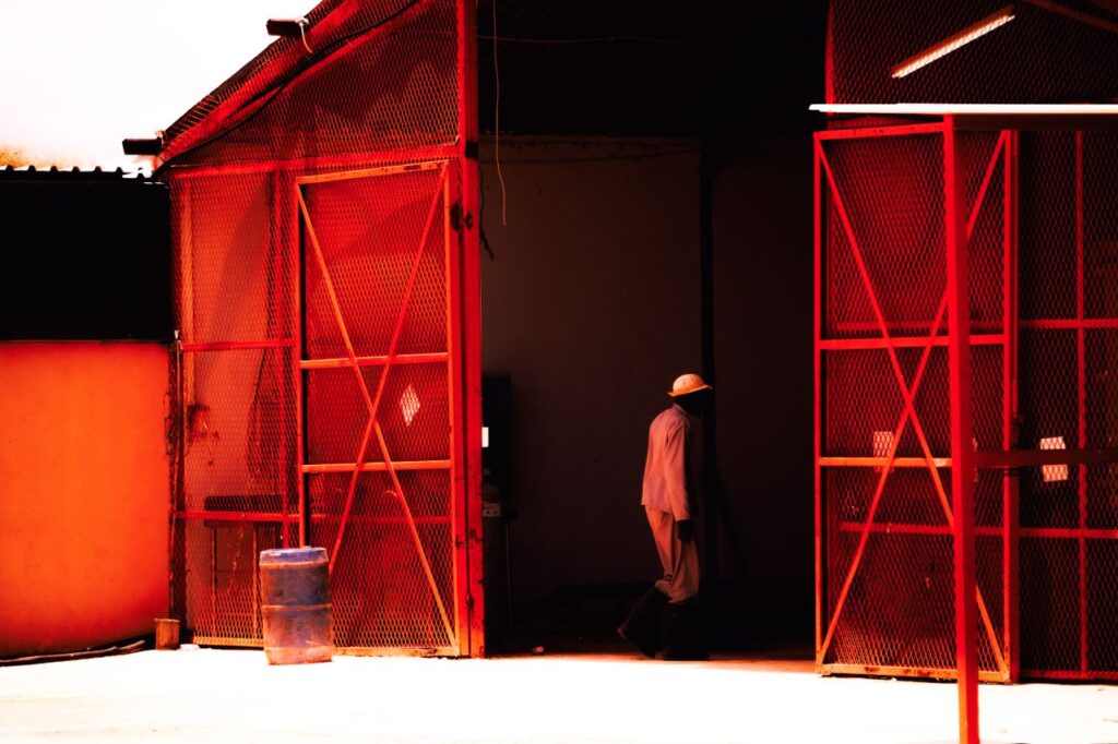 A man enters a vividly colored red warehouse, casting dynamic shadows from the sunlight outside.