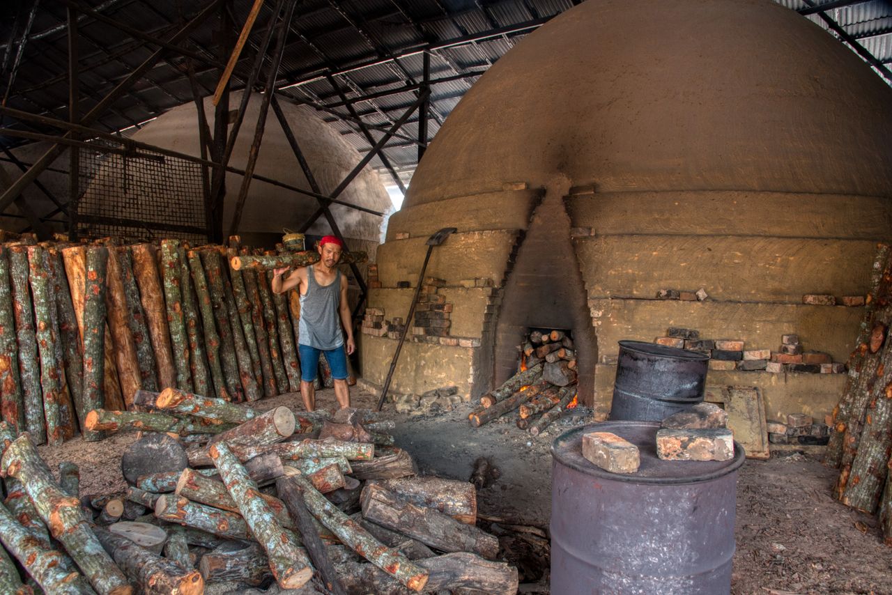 A laborer organizes logs near an igloo-shaped kiln for charcoal production in an industrial setting.